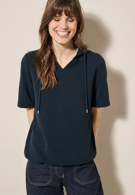 Lächelnde Frau mit dunkelblauem Shirt und kurzen Ärmeln vor hellem Hintergrund.